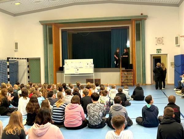 Schüler sitzen in der Turnhalle auf dem Boden vor einer Bühne.